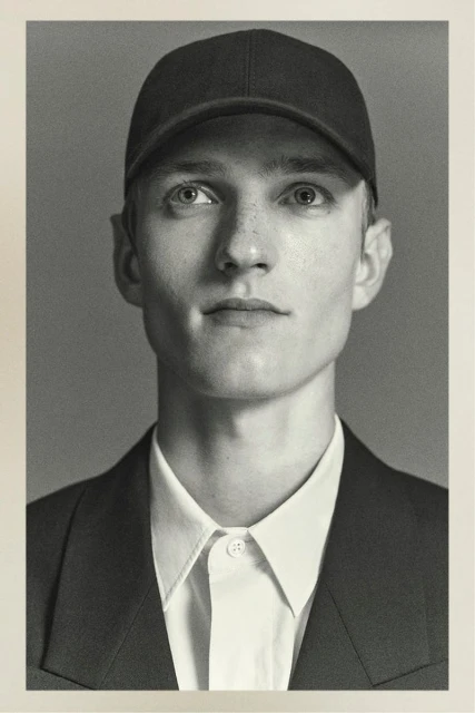 Portrait of a young fair-skinned adult male wearing a baseball cap, collared shirt, and blazer, set against a muted grey backdrop.
