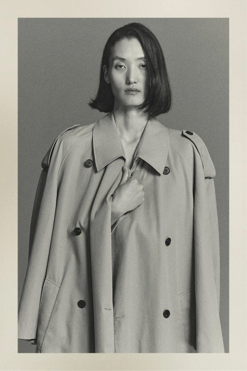 Studio shot of an Asian woman in a light beige trench coat looking at the camera with a neutral expression in portrait orientation.