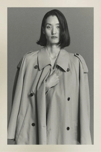 Studio shot of an Asian woman in a light beige trench coat looking at the camera with a neutral expression in portrait orientation.
