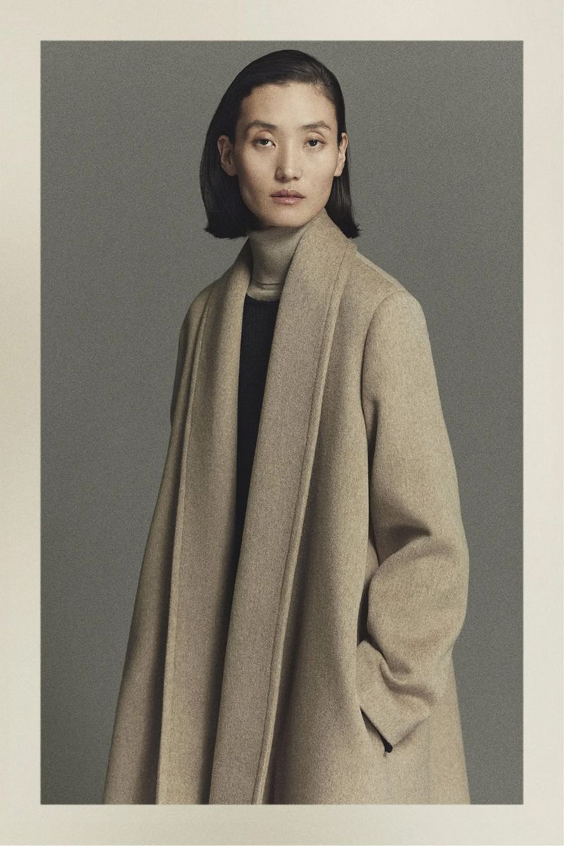 Elegant studio portrait of a woman in a tan overcoat looking at the camera against a grey backdrop, showcasing fashion and style.