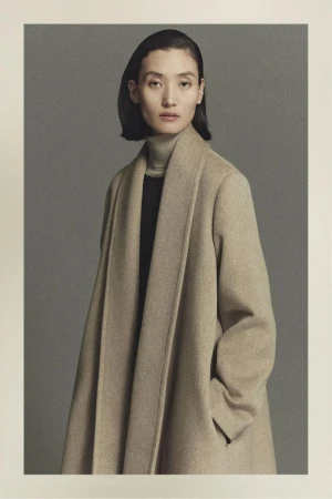 Elegant studio portrait of a woman in a tan overcoat looking at the camera against a grey backdrop, showcasing fashion and style.
