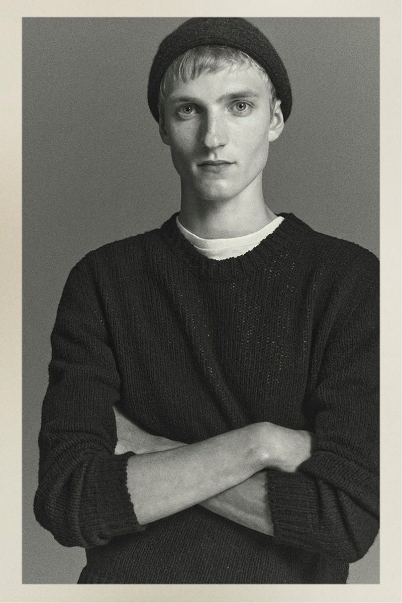 Portrait of a fair-skinned young man in a knit hat and sweater, posing with arms crossed against a solid gray background.