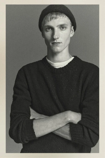 Portrait of a fair-skinned young man in a knit hat and sweater, posing with arms crossed against a solid gray background.
