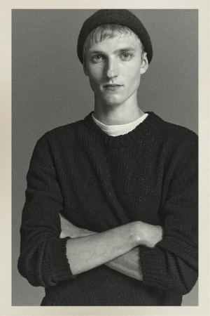 Portrait of a fair-skinned young man in a knit hat and sweater, posing with arms crossed against a solid gray background.