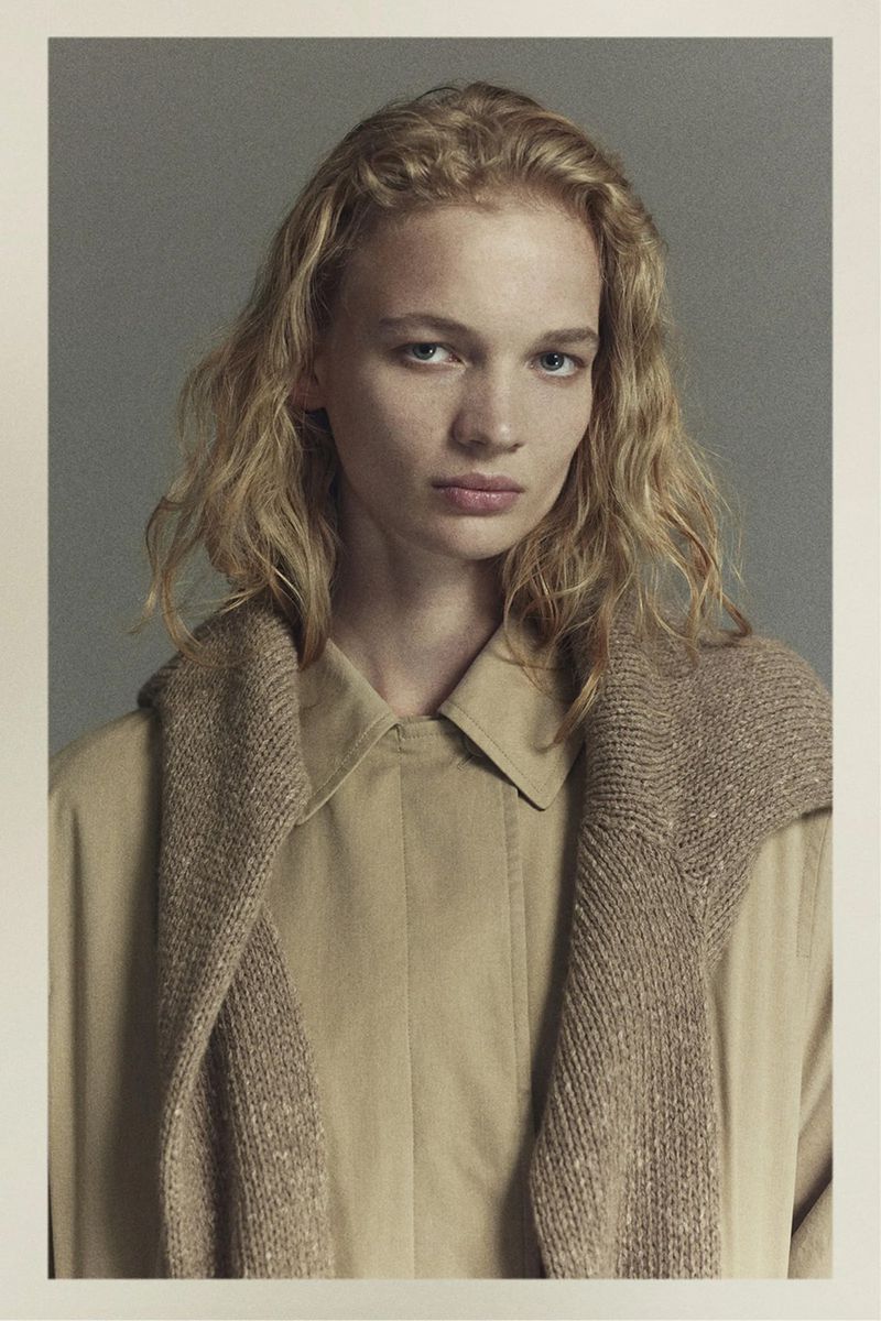 Portrait of a fair-skinned woman with blonde wavy hair wearing a tan shirt and sweater, gazing directly at the camera in a studio shot.