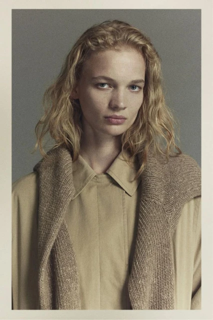 Portrait of a fair-skinned woman with blonde wavy hair wearing a tan shirt and sweater, gazing directly at the camera in a studio shot.