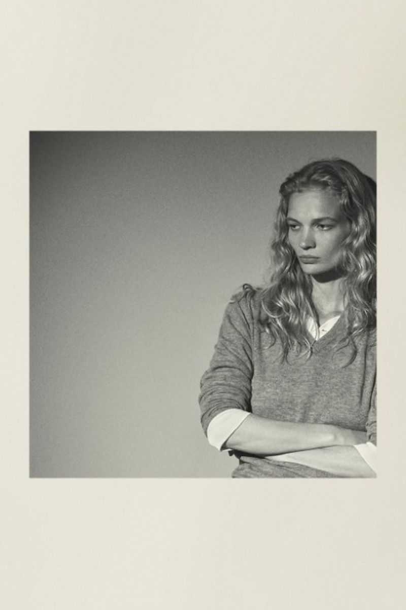 Monochrome portrait of a woman with blonde wavy hair wearing a grey sweater, arms crossed, looking away with a neutral expression.