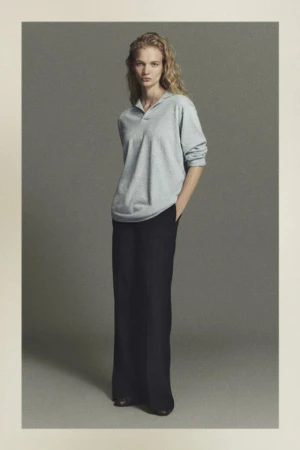Fashion shot of a woman with blonde curly hair wearing a light blue long-sleeved polo shirt and black pants in a studio setting.