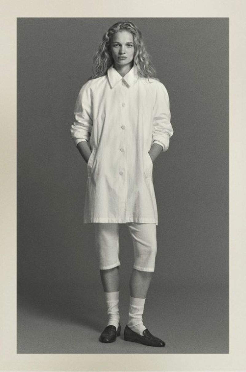 Full-length studio shot of a blonde woman in white coat, shorts and socks, with dark loafers, posing against a grey backdrop.