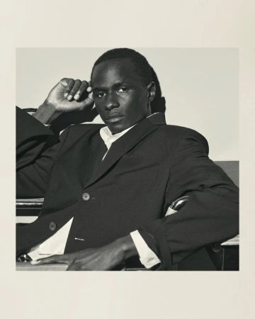 Portrait of a Black man in a suit, seated with hand on head, exuding confidence in a photo studio with soft, diffused lighting.