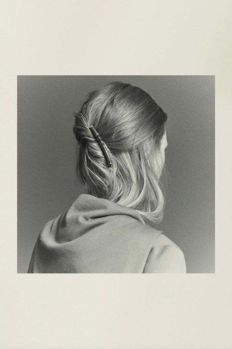 Blond woman with a twist hairstyle, wearing a top, shot from the back. Focus on the hair accessory, against a grey backdrop.
