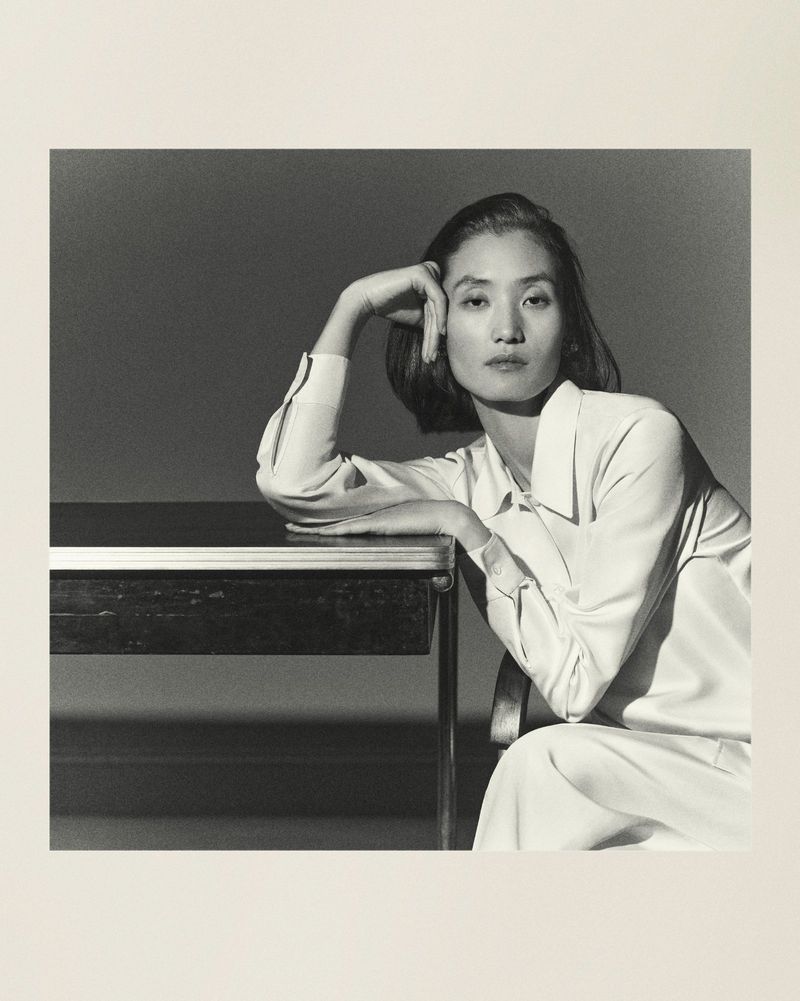Monochrome portrait of an Asian woman in a light-colored, button-down shirt, seated at a dark table in a photo studio.