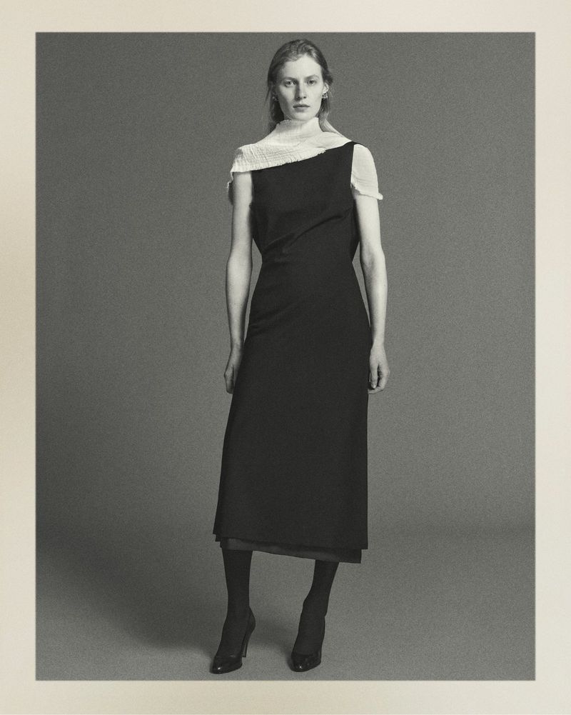 Full-length studio shot of a woman in a black dress with sheer white fabric draped over one shoulder, wearing sheer tights and high heels.