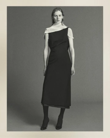 Full-length studio shot of a woman in a black dress with sheer white fabric draped over one shoulder, wearing sheer tights and high heels.