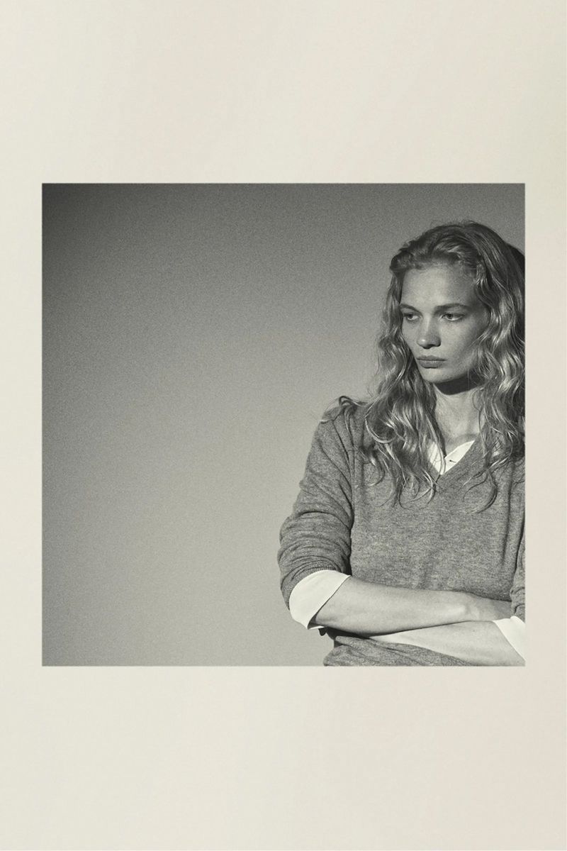 Portrait of a fair-skinned adult woman with long blonde hair, wearing a gray sweater, and arms crossed against a plain background.
