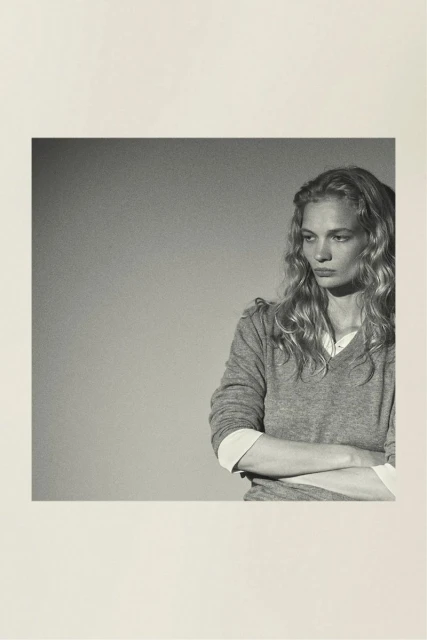 Portrait of a fair-skinned adult woman with long blonde hair, wearing a gray sweater, and arms crossed against a plain background.