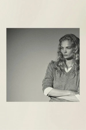 Portrait of a fair-skinned adult woman with long blonde hair, wearing a gray sweater, and arms crossed against a plain background.