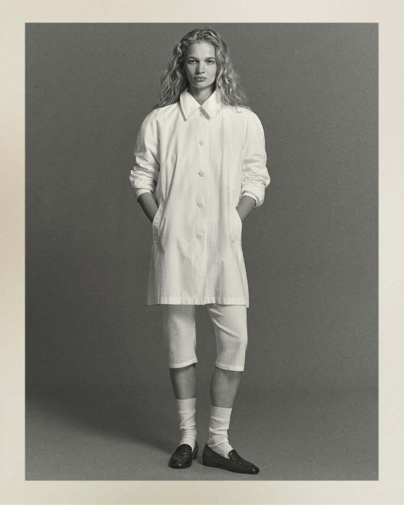 Full shot of a young, blonde female fashion model in a white tunic, culottes, and loafers posing in a photo studio against a grey background.