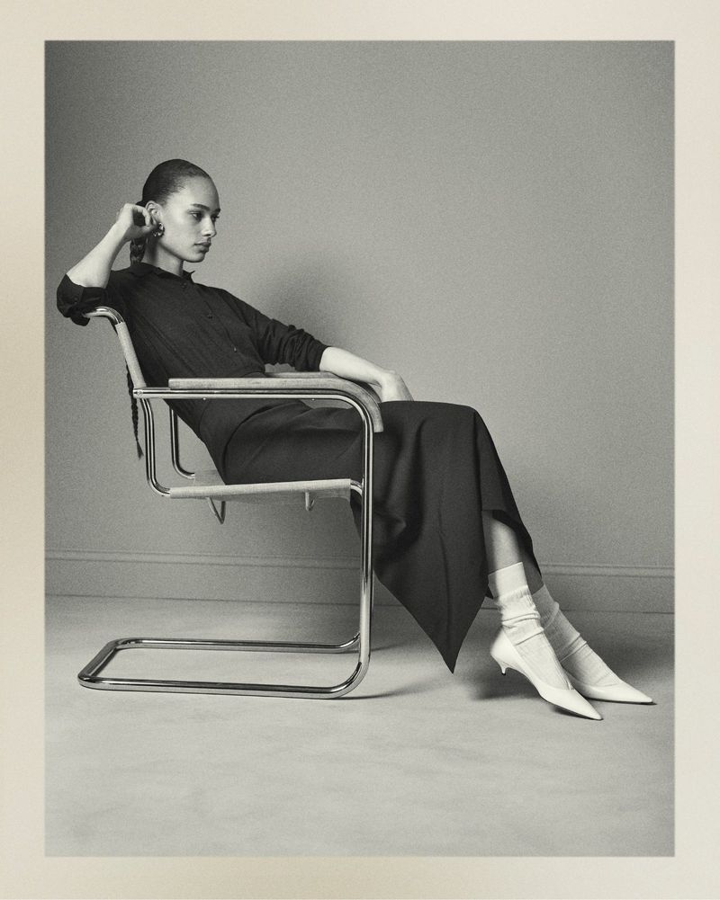 Black and white portrait of a seated woman in dark, full skirt, light socks and heels, posing in a chrome and wood chair.