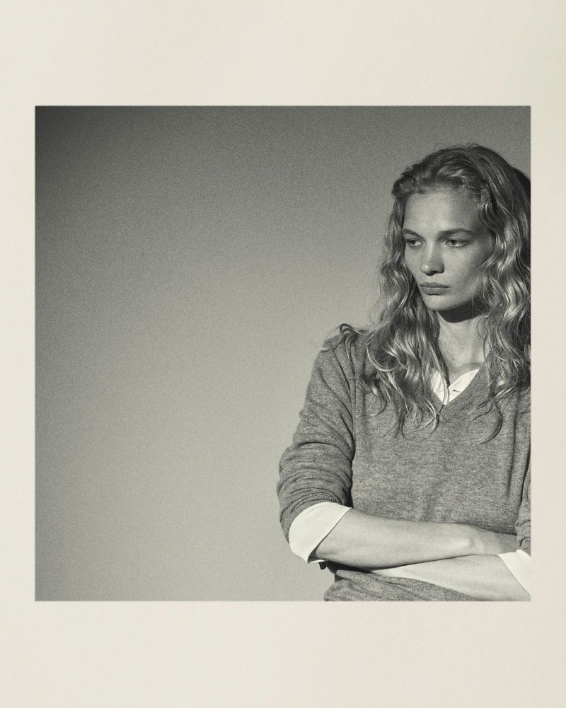 Portrait of a blonde woman in a grey sweater with crossed arms. Studio-style with a minimalist background and neutral colour palette.