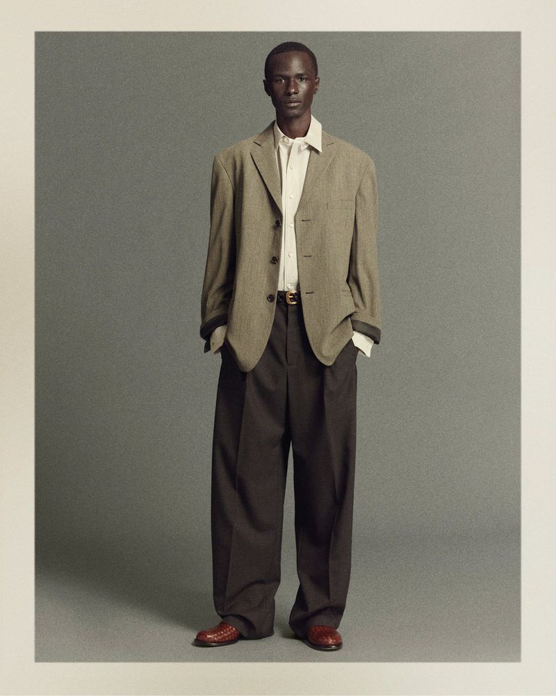 Portrait of a dark-skinned male model wearing an olive-green blazer, beige dress shirt, oversized brown trousers, and brown leather shoes.