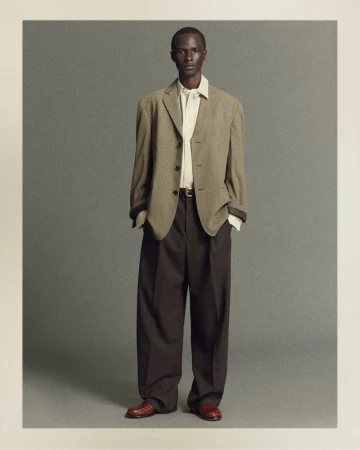 Portrait of a dark-skinned male model wearing an olive-green blazer, beige dress shirt, oversized brown trousers, and brown leather shoes.