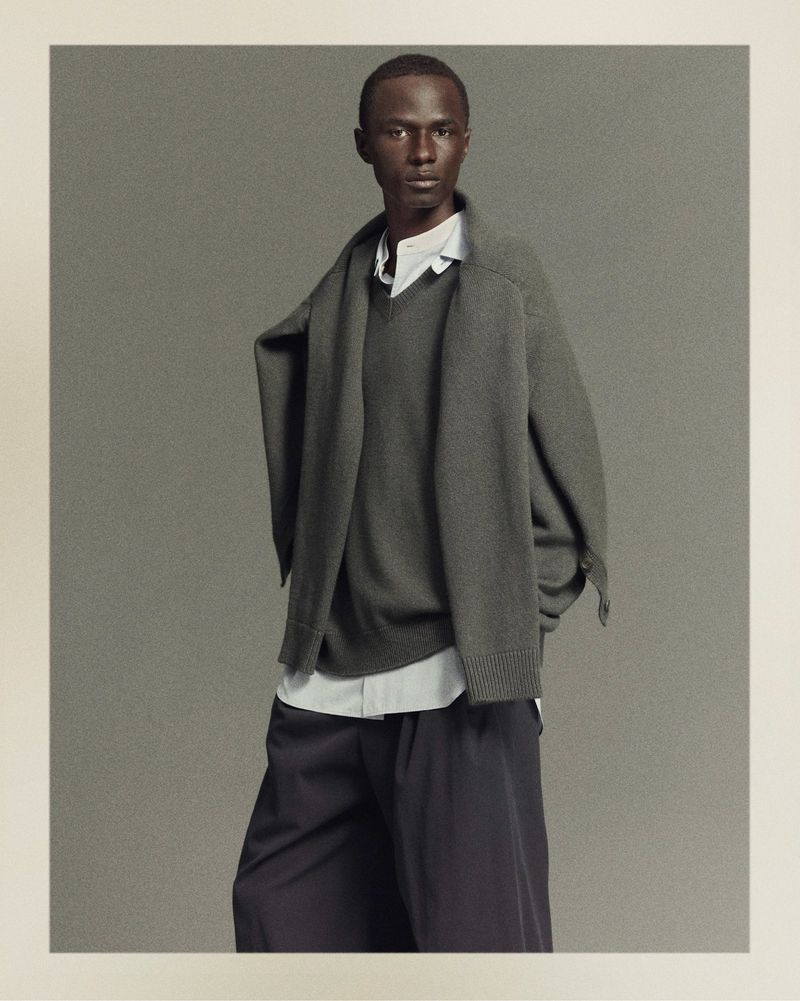 Fashion shot of an adult Black male in a grey sweater, white button-down shirt, and black pants, posing indoors against a neutral backdrop.