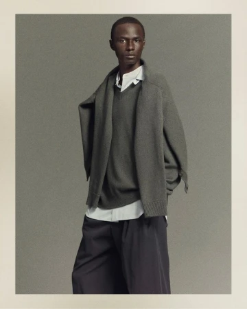 Fashion shot of an adult Black male in a grey sweater, white button-down shirt, and black pants, posing indoors against a neutral backdrop.