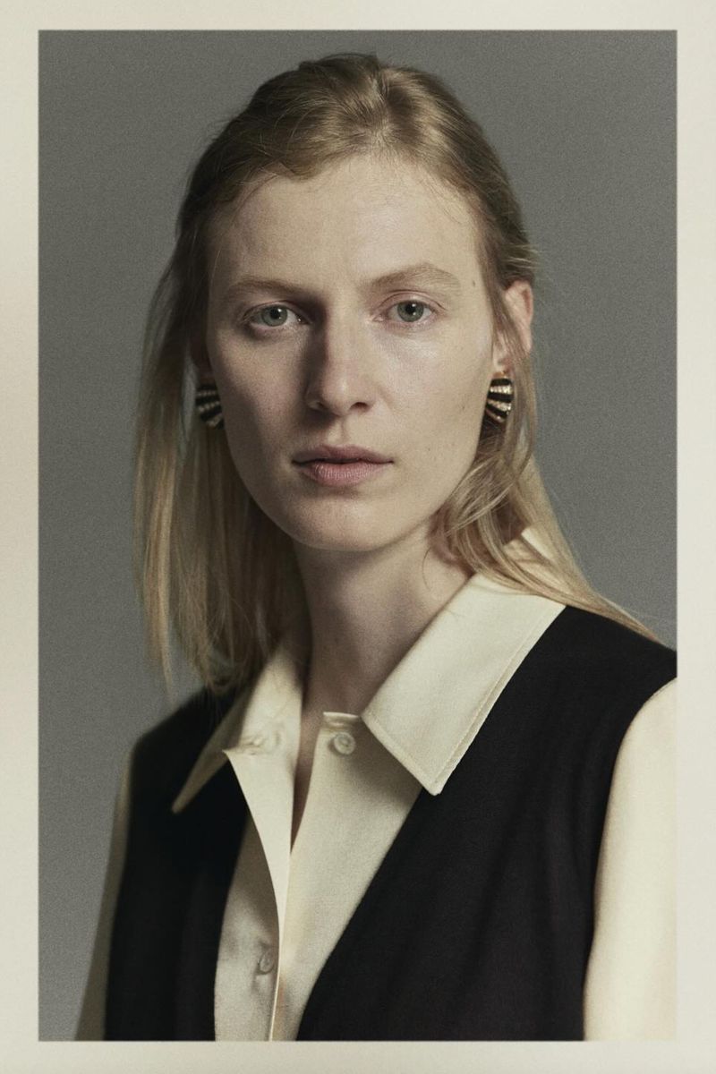 Portrait of a blonde woman in a black and cream colored top, wearing black and white striped earrings, against a grey backdrop.