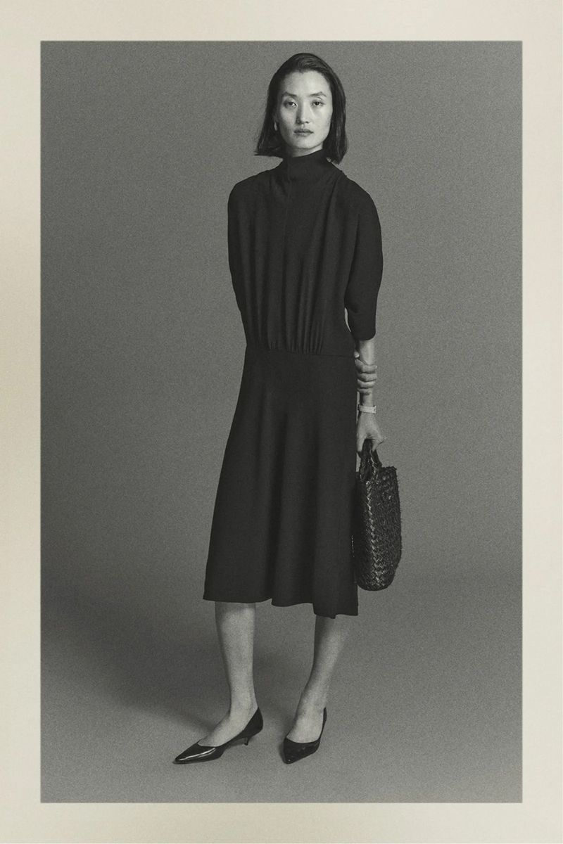 Monochrome portrait of a woman in an elegant black dress, holding a woven handbag, standing against a muted grey backdrop in a photo studio.