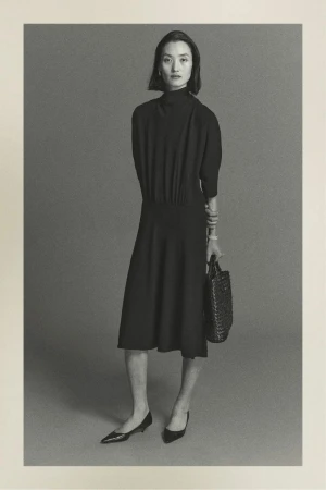 Monochrome portrait of a woman in an elegant black dress, holding a woven handbag, standing against a muted grey backdrop in a photo studio.