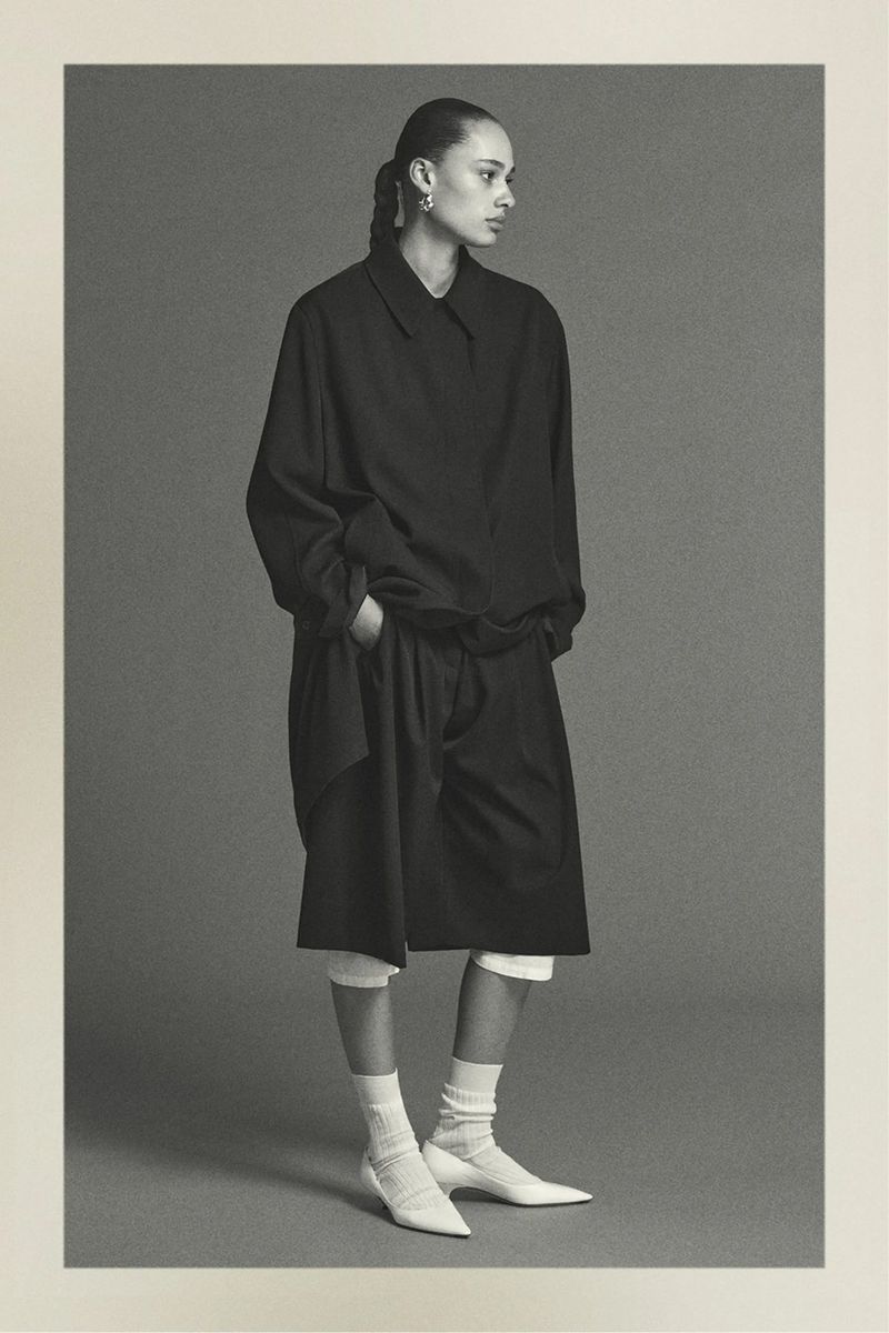 A black-and-white fashion shot of a young adult woman in a black shirt, shorts, and white socks and shoes, set against a neutral background.