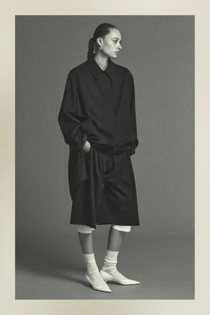A black-and-white fashion shot of a young adult woman in a black shirt, shorts, and white socks and shoes, set against a neutral background.