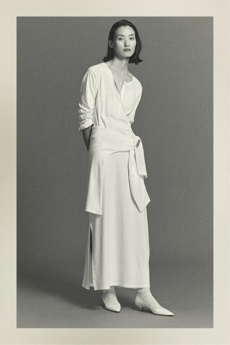Elegant Asian woman posing in a long white wrap dress with tie detail, paired with white kitten heels, against a neutral gray studio backdrop.