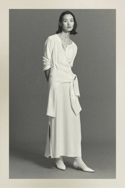 Elegant Asian woman posing in a long white wrap dress with tie detail, paired with white kitten heels, against a neutral gray studio backdrop.
