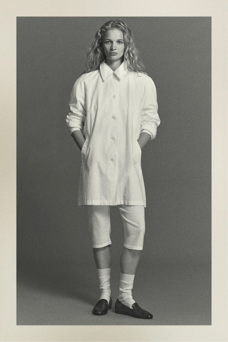 Monochrome portrait of a blonde woman in a white coat, capri pants, socks, and loafers, standing indoors against a grey background.