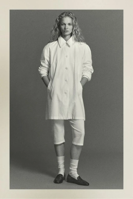 Monochrome portrait of a blonde woman in a white coat, capri pants, socks, and loafers, standing indoors against a grey background.