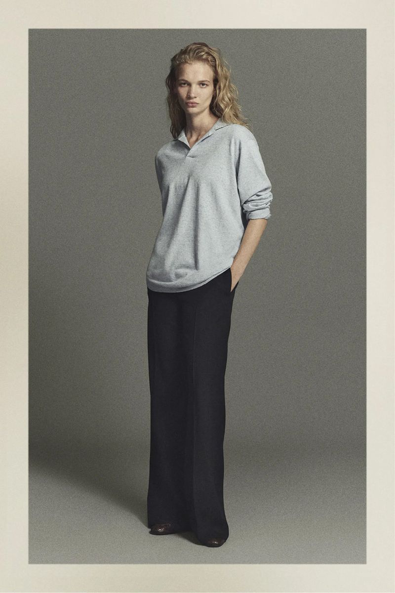 Full-length shot of an adult female model in a light grey shirt and dark trousers in a neutral-toned studio setting.