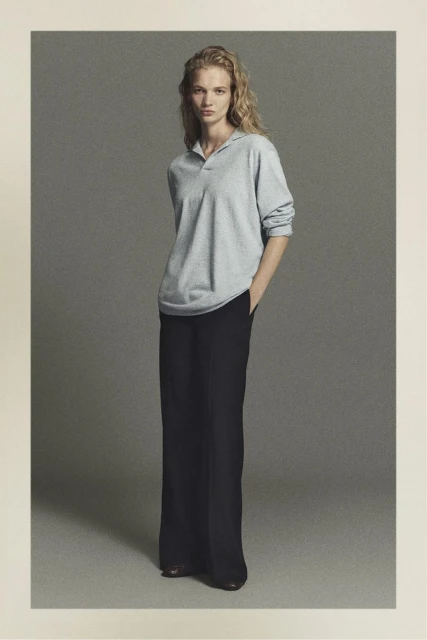 Full-length shot of an adult female model in a light grey shirt and dark trousers in a neutral-toned studio setting.
