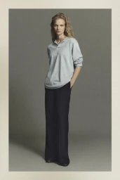 Model standing in a light grey polo shirt and black trousers against a neutral grey studio background