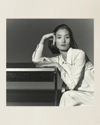 Model in a white collared shirt resting her elbow on a desk against a grey studio backdrop.