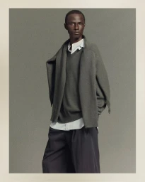 Model Malick Bodian posing in a layered grey V-neck sweater, draped knit cardigan, and dark trousers against a grey backdrop.