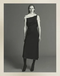 Model standing against a neutral studio backdrop wearing a dark sleeveless midi dress with a textured white scarf