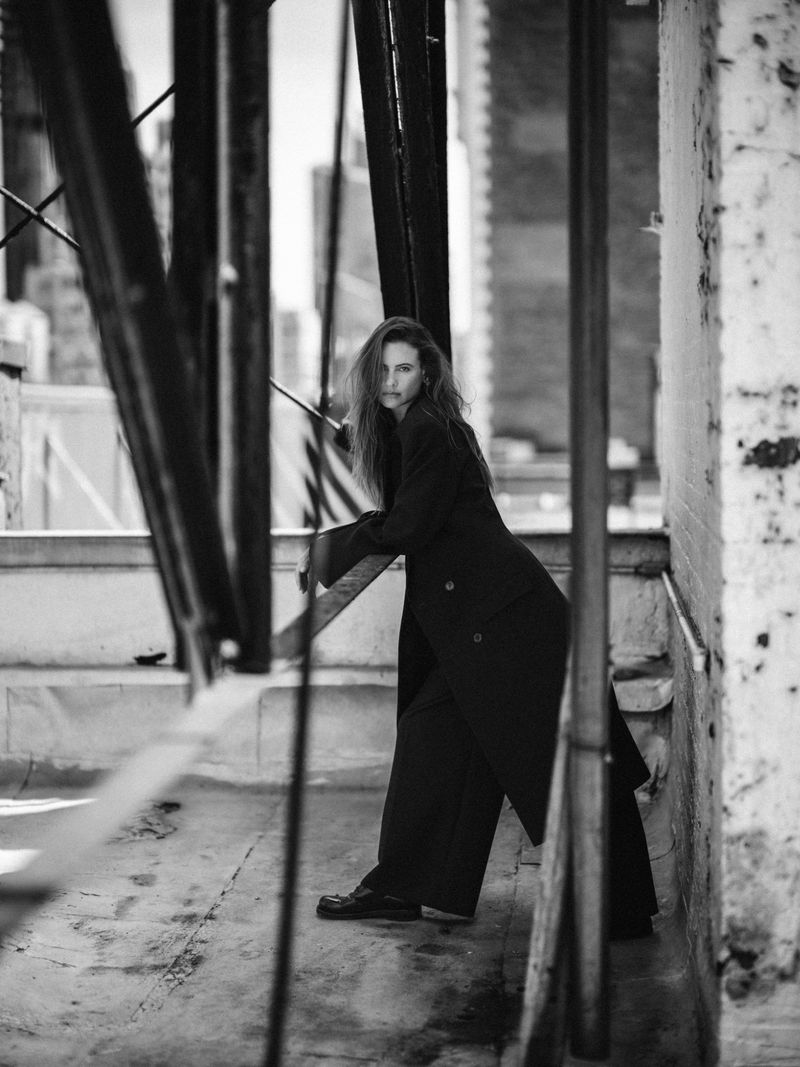 Model Behati Prinsloo posing in an oversized long dark coat and trousers against metal rooftop beams in black and white.