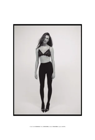Full-length black and white portrait of Behati Prinsloo wearing a black lace bralette, black tights, and heels on a white backdrop.
