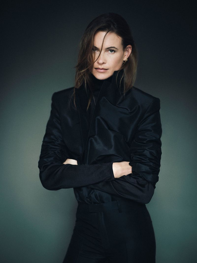 Model Behati Prinsloo wearing a black blazer and turtleneck with arms crossed against a dark muted background.