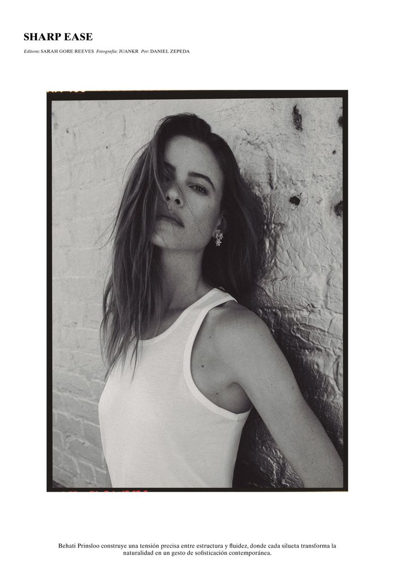 Black and white editorial portrait of Behati Prinsloo in a white tank top leaning against a textured painted brick wall.