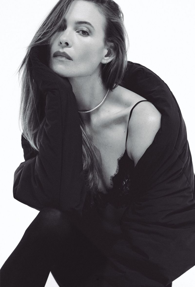 Monochrome portrait of model Behati Prinsloo wearing an oversized black coat and delicate choker necklace against a white background.