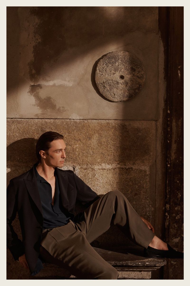 A stylish adult male poses with a jacket and brown pants next to a concrete wall with a stone decoration in soft, natural light.