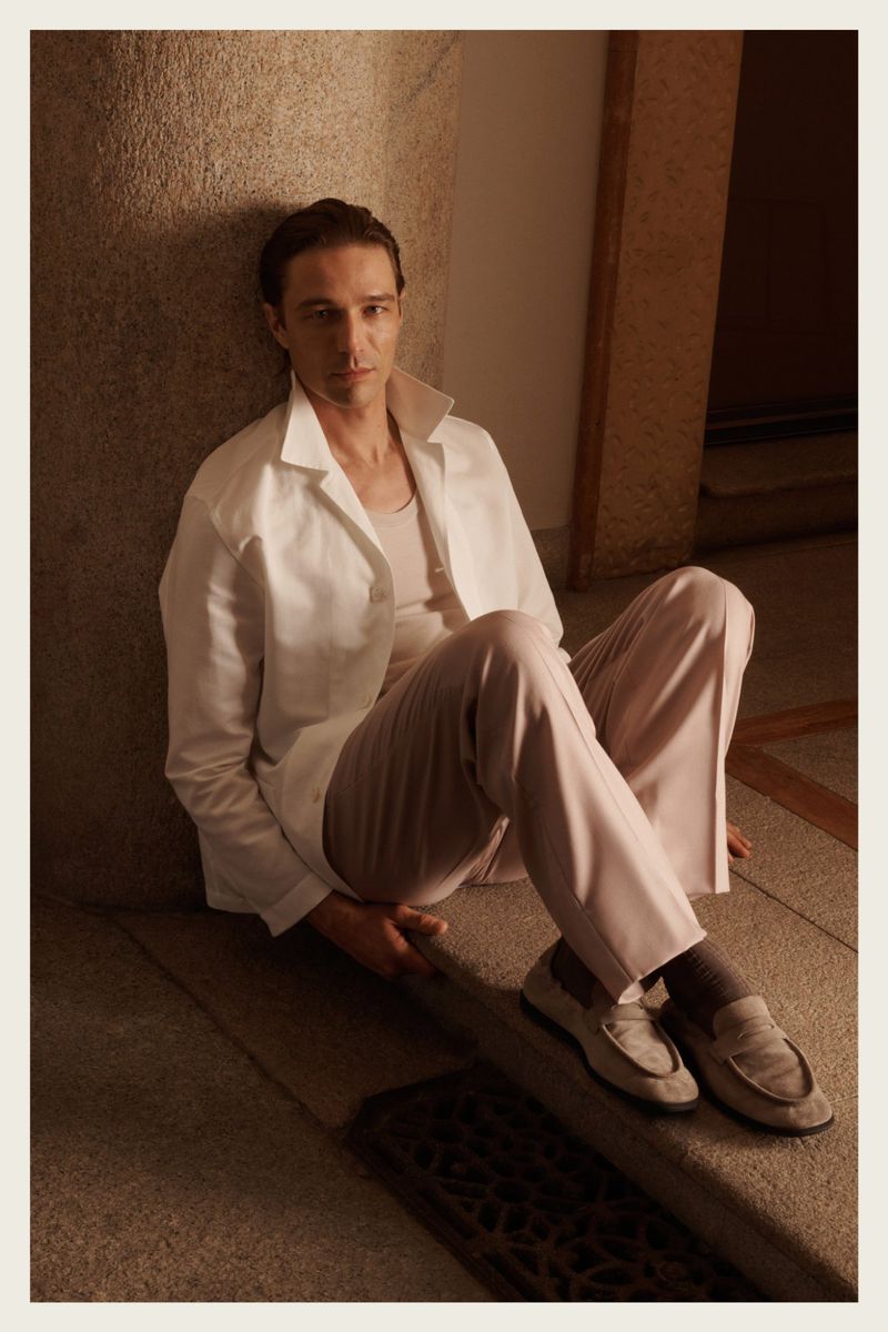 A fair-skinned man in a white shirt and beige pants is leaning against a wall, showcasing indoor fashion with loafers in soft, warm lighting.